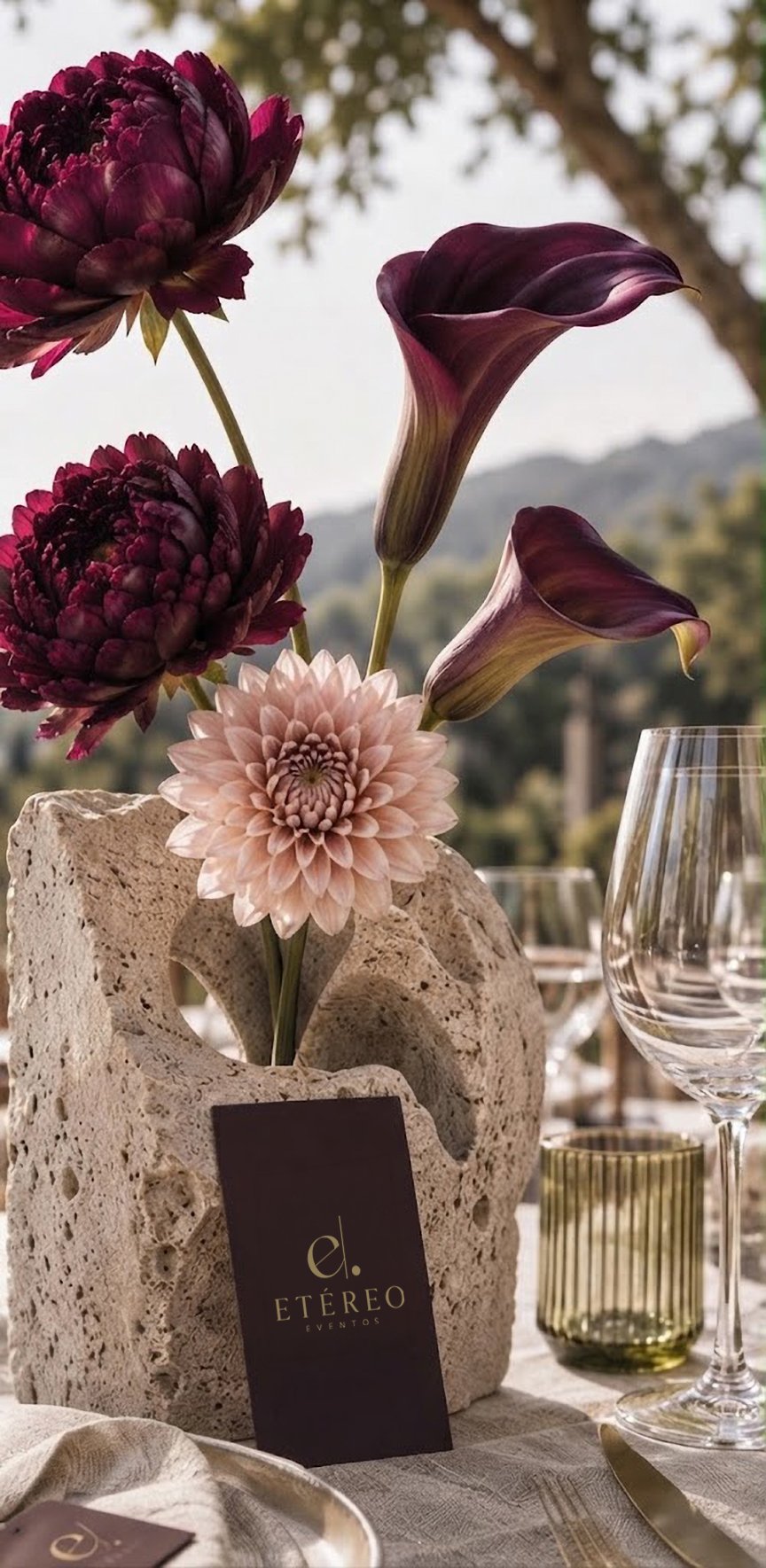 Peonías color vino, alcatraces y una dalia rosa en un florero de piedra sobre una mesa elegante